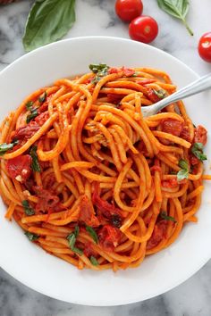 a white bowl filled with pasta and tomatoes on top of a marble table next to fresh basil