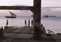 several people standing near the water with bikes and boats in the background at sunset or dawn