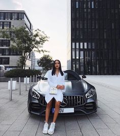 a woman sitting on the hood of a sports car in front of tall black buildings