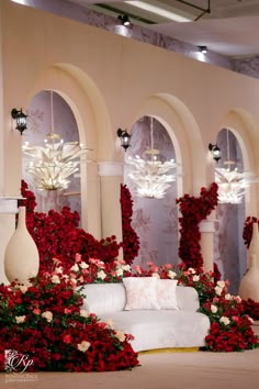 a white couch surrounded by red flowers and chandeliers in a room with arches