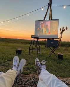 two people are sitting in the grass watching tv with their feet propped up on a blanket