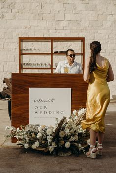 a woman in a yellow dress standing next to a man behind a bar with flowers on it