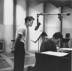 two men are standing in front of a piano and microphone, one is looking at the other