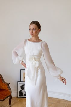 a woman wearing a white dress with long sleeves and a bow at the waist, standing on a wooden floor