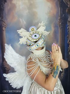 a woman wearing a white mask with wings and pearls on her face, standing in front of a painting