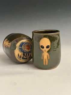 two ceramic cups with designs on them sitting next to each other, one has an alien face painted on it