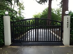 an iron gate with two white pillars and a black fence on the other side, in front of some trees