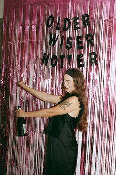 a woman in a black dress standing next to a pink curtain and holding a wine bottle