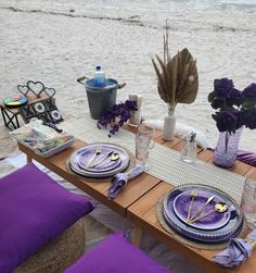 the table is set with purple flowers and place settings for two people on the beach