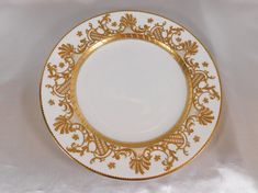 a white and gold plate with an ornate design on the rim, sitting on a white table cloth