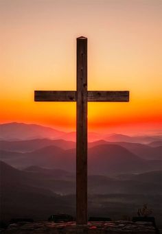 a wooden cross on top of a hill with mountains in the background at sunset or sunrise