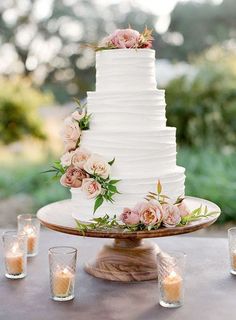 a white wedding cake with pink flowers and greenery sits on a table surrounded by candles