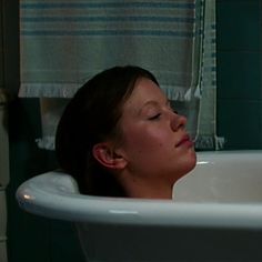 a woman taking a bath in a white sink with her eyes closed and head resting on the faucet