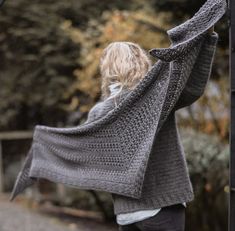 a woman is standing outside with her arms in the air and she has a gray knitted shawl over her shoulders