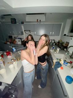 two young women standing in a kitchen, one is covering her face with her hands