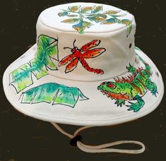 This is a great hat for the outdoors! Whether you're into gardening, fishing, or walking on the beach, this hat will shield you from that summer sun. Adjustable chin cord will keep it from blowing away on a windy day. 100% heavy cotton canvas, natural color; individually hand painted with a green iguana, various tropical foliage, and a dragonfly! Each hat is painted freehand, so no two look exactly alike. The acrylic paints used are permanent, so if your hat happens to land in the water, not to worry. The hat might be all wet, but the painted design will still be there. One size, fits L-XL. Hand wash. Bucket Hat Painting, Animal Bucket Hat, Hat Painting, Painted Shirt, Green Iguana, Painted Clothing, Outback Hat, Hand Painted Clothing, Winston Salem Nc