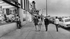 black and white photograph of people walking down the sidewalk in front of a storefront