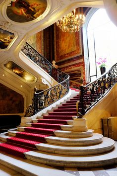 a staircase with red carpeted steps and chandelier in the background, along with paintings on the walls