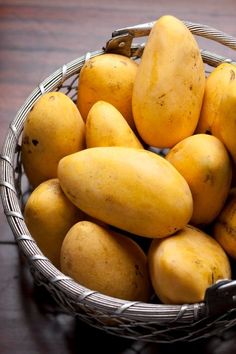 a basket full of yellow mangoes on a wooden table with other fruit in the background