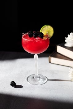 a red cocktail garnished with blackberries and lime sits on a table next to an open book