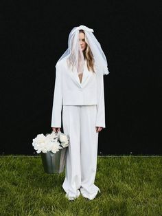 a woman in a white suit and veil holding a bucket with flowers on the grass