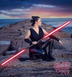 a woman is sitting on the rocks with two lightsabes in her hand and one light saber across her leg