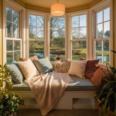 a window seat with many pillows on it in front of a large bay window filled with plants