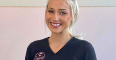 a woman with blonde hair wearing a black shirt and smiling at the camera while standing in front of a white wall