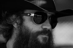 a close up of a person wearing sunglasses and a hat with a long beard in black and white