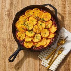 a skillet filled with potatoes on top of a wooden table next to silverware