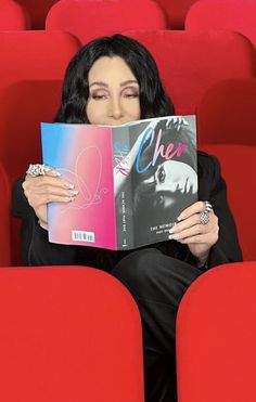 a woman sitting in a red chair holding up a book to her face and looking at the camera