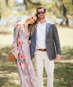 a man and woman standing next to each other in the grass with trees behind them