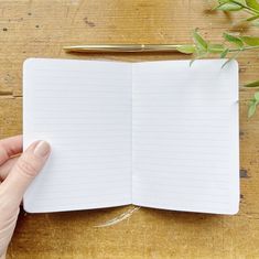 a hand holding a pen over an open notebook on a wooden table next to a plant