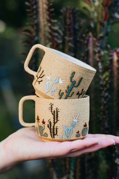 two coffee mugs with cactus designs on them are held in front of a plant