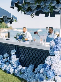 two people sitting at a bar with blue hydrangeas on the side and an umbrella over them