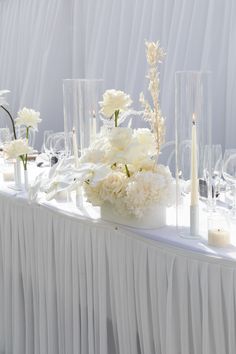 the table is set with white flowers, candles and glass vases on top of it