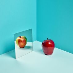 an apple sitting next to a mirror on top of a white table with a blue wall in the background