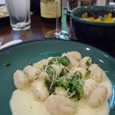 a blue plate topped with dumplings covered in cheese and greens next to a glass of wine