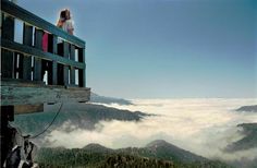 a woman sitting on top of a wooden bench looking out over the clouds and mountains