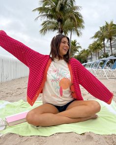 a woman sitting on top of a beach next to a palm tree and wearing a pink cardigan