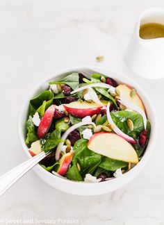 a white bowl filled with spinach, apples and cranberries next to a cup of tea