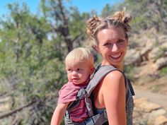 a woman carrying a baby in a carrier on top of a rock covered hillside with trees behind her