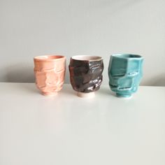 three different colored cups sitting on top of a white table next to each other in front of a wall