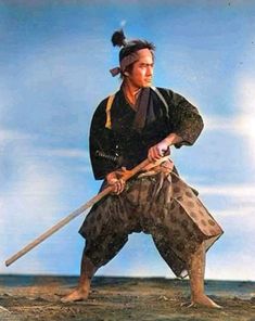 a man holding two swords while standing on top of a field next to the ocean