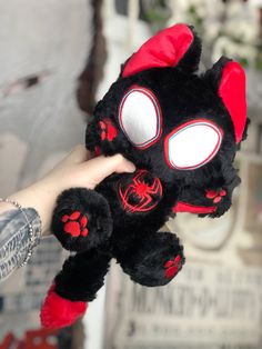 a hand holding a black and red stuffed animal with white eyes, claws and paws