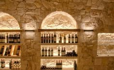 several shelves filled with bottles of wine in a stone walled room, lit by lights