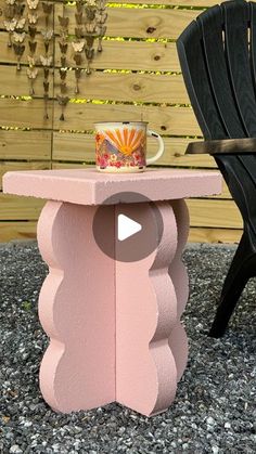 a pink table with a coffee cup on it next to a black chair and wooden fence