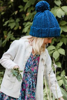 a woman wearing a blue knitted hat while walking through the woods with flowers in her hand