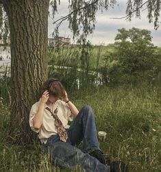 a man sitting in the grass next to a tree talking on a cell phone while holding his head