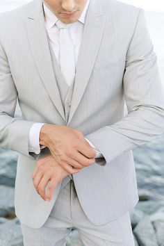 a man in a gray suit and white shirt is standing by the water with his hands on his lapel